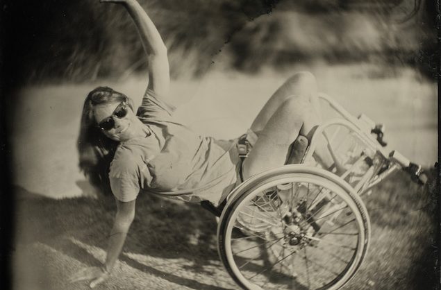 La patineuse Tracie Gargacochea effectue une figure d'équilibre en fauteuil roulant, les yeux masqués par des lunettes de soleil et le visage tourné vers l'appareil photo.