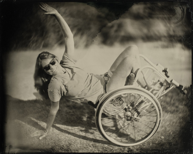 La patineuse Tracie Gargacochea effectue une figure d'équilibre en fauteuil roulant, les yeux masqués par des lunettes de soleil et le visage tourné vers l'appareil photo.