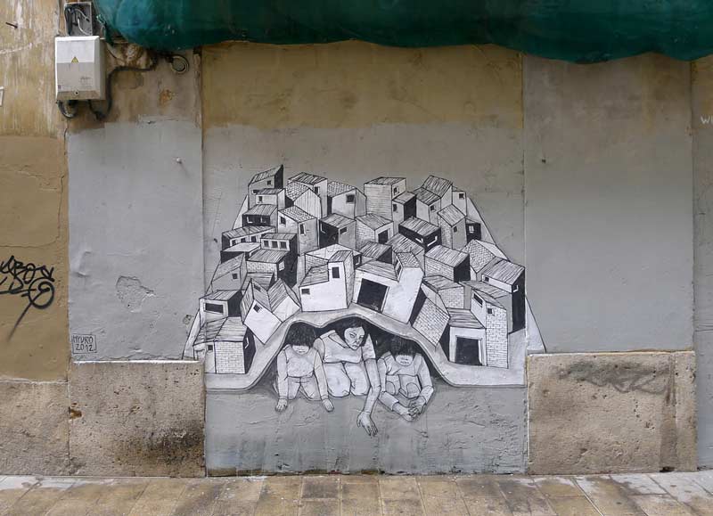 Vue d'un mur extérieur peint d'une fresque représentant une ville en noir et blanc. La ville se soulève comme un tapis et dessous 3 silhouettes humaines sont comme écrasées par le poids de la ville.